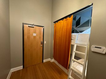 Cozy budget room with wooden entry door, bamboo floors, white bunk bed ladder and bright orange curtain revealing a top bunk and geometric accent wall.