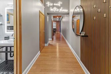 Stylish modern loft hallway with light wood floors and gray walls, neon sign above a doorway, round mirror on a slatted wood accent wall, bathroom sink visible on the left and exposed brick room at the far end.