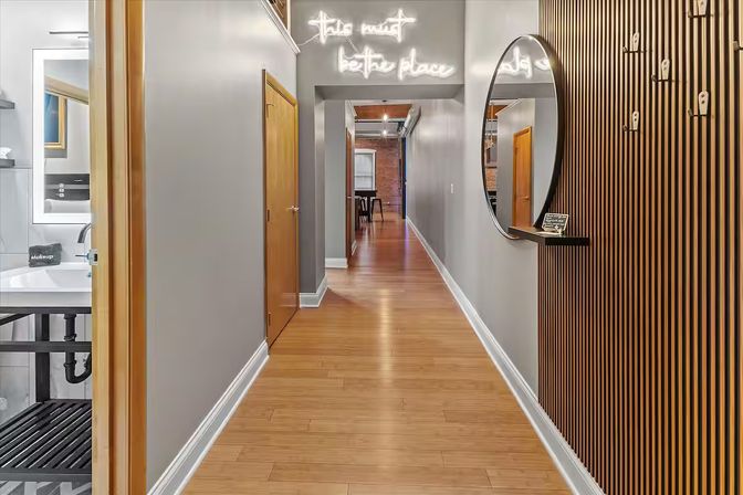Stylish modern loft hallway with light wood floors and gray walls, neon sign above a doorway, round mirror on a slatted wood accent wall, bathroom sink visible on the left and exposed brick room at the far end.