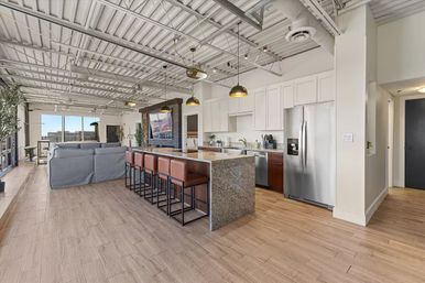 Open-concept urban loft apartment with granite kitchen island and six leather barstools, stainless steel appliances, white cabinetry, exposed industrial ceiling, wood floors, pendant lights and floor-to-ceiling windows with city skyline views.