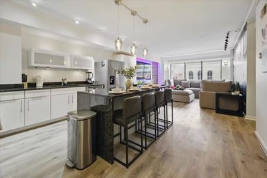 Modern open-concept apartment kitchen and living room with black granite island and four bar stools, stainless steel appliances, pendant lights, light hardwood floors, and a gray sectional by large windows with subtle purple accent lighting.