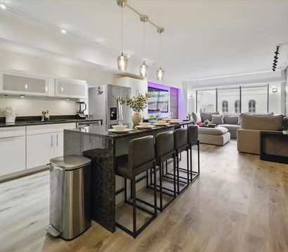 Modern open-concept apartment kitchen and living room with black granite island and four bar stools, stainless steel appliances, pendant lights, light hardwood floors, and a gray sectional by large windows with subtle purple accent lighting.