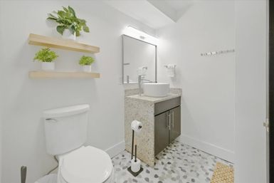 Bright minimalist powder room with white walls, vessel sink on a speckled stone vanity and dark cabinet, wall mirror and chrome faucet, floating wooden shelves with potted green plants, white toilet and pebble-pattern floor tiles.