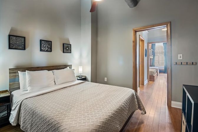 Modern urban loft bedroom with a large bed in white quilted bedding, wood headboard, three black framed wall art pieces, nightstand with lamp, and hardwood floors leading through an open doorway to another room.
