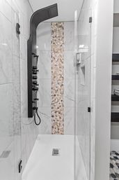 Sleek walk-in shower with matte black multi-jet shower panel, vertical pebble-mosaic accent strip, white marble-look wall tiles and frameless glass door in a modern bathroom.