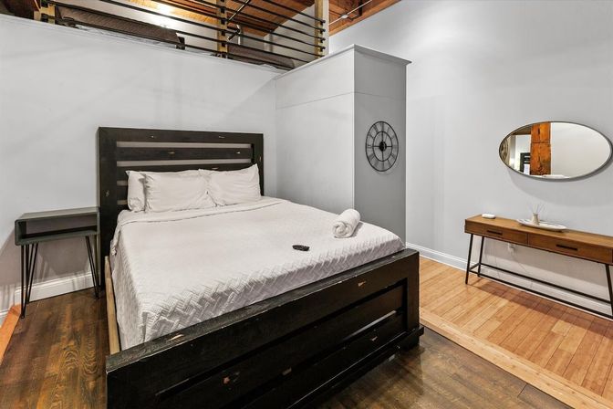 Modern loft bedroom interior with dark wood platform bed, crisp white bedding and rolled towel, wall clock, oval mirror, wooden floors and visible loft railing above.