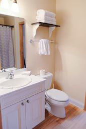 Cozy small beige bathroom with white vanity and sink, toilet, chrome fixtures, stacked white towels on a wall shelf, towel bar, wood-look floor and patterned shower curtain visible in mirror
