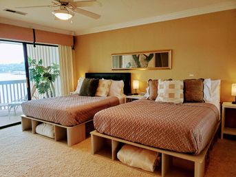 Cozy lakefront bedroom with two double beds, warm beige decor, sliding balcony door with water view, bedside lamps and ceiling fan.