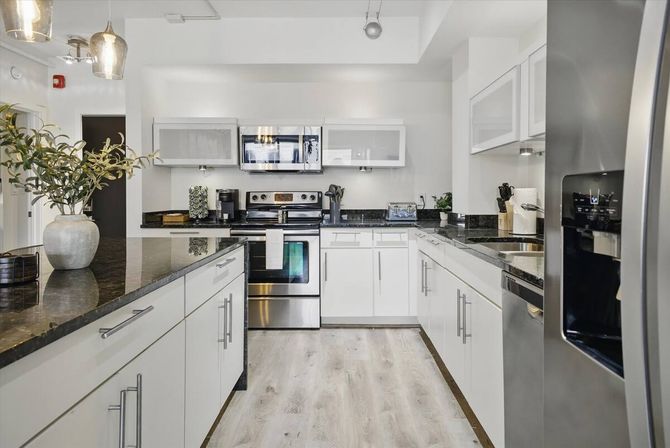 Bright, modern white U-shaped kitchen with stainless steel appliances, black granite countertops, island with potted plant and light wood floors