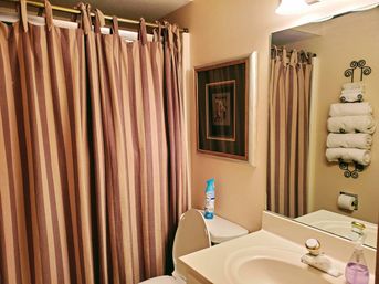 Cozy residential bathroom with brown-and-beige striped shower curtain, white toilet topped with an air freshener, cream sink and mirror, and a wall-mounted wrought-iron rack holding rolled white towels.