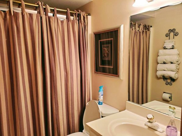 Cozy residential bathroom with brown-and-beige striped shower curtain, white toilet topped with an air freshener, cream sink and mirror, and a wall-mounted wrought-iron rack holding rolled white towels.