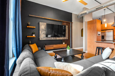 Modern urban loft living room with dark accent wall and wall-mounted flat-screen TV, gray sectional sofa with mustard pillows, black coffee table, exposed ductwork, blue curtains and open kitchen with wooden cabinets and stainless refrigerator.