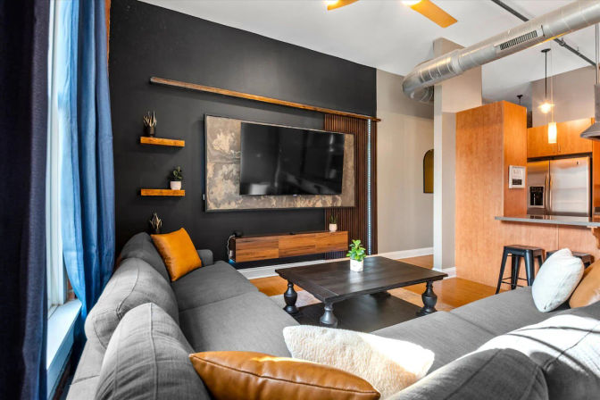 Modern urban loft living room with dark accent wall and wall-mounted flat-screen TV, gray sectional sofa with mustard pillows, black coffee table, exposed ductwork, blue curtains and open kitchen with wooden cabinets and stainless refrigerator.