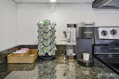Keurig single-serve coffee station on a glossy dark granite kitchen countertop with a rotating K-Cup holder, water reservoir, and wicker tray of sweeteners — ready for the morning brew.
