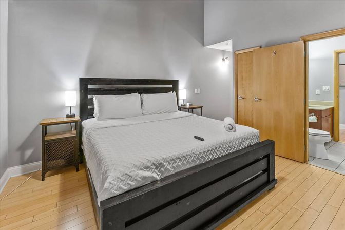 Contemporary bedroom with king-size dark wood bed, crisp white bedding, bedside tables with lamps, light hardwood floors and open door to an ensuite bathroom.