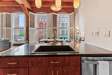 Urban loft kitchen island with black sink and polished countertop, exposed wooden beams and plantation shutters framing tall arched windows overlooking red-brick historic downtown buildings.