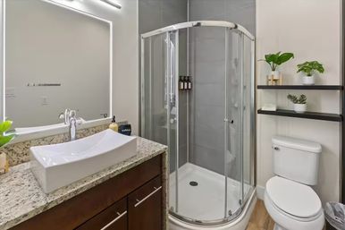 Bright modern bathroom with curved glass corner shower, white rectangular vessel sink on a granite countertop, dark wood vanity, backlit mirror, toilet and black floating shelves with potted plants on neutral gray tile and wood-look floor.