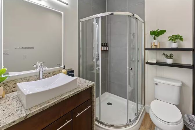 Bright modern bathroom with curved glass corner shower, white rectangular vessel sink on a granite countertop, dark wood vanity, backlit mirror, toilet and black floating shelves with potted plants on neutral gray tile and wood-look floor.