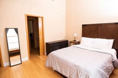 Cozy bright bedroom with dark wooden headboard, white bedding, hardwood floors, black dresser, leaning full-length mirror, bedside lamp and open sliding door.