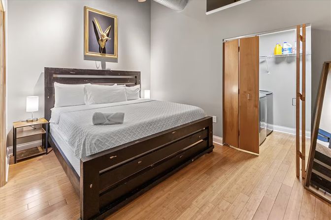 Modern loft-style bedroom with king-size bed and white linens, dark wood frame, bedside lamp, framed animal skull artwork, bamboo hardwood floors, and an open closet showing washer/dryer and shelving.