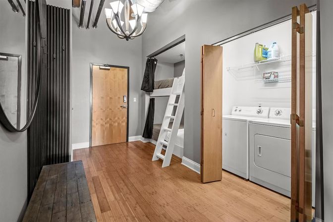 Modern urban apartment entryway with hardwood floors, wooden entry door, built-in white bunk bed and ladder, open laundry closet with washer and dryer, gray walls and round mirror