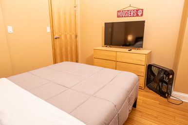 Cozy tan bedroom with a neatly made bed, light wood dresser with flat-screen TV, box fan on hardwood floor and a red 'Hoosiers' wall sign.