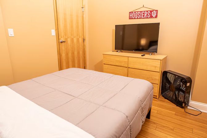 Cozy tan bedroom with a neatly made bed, light wood dresser with flat-screen TV, box fan on hardwood floor and a red 'Hoosiers' wall sign.