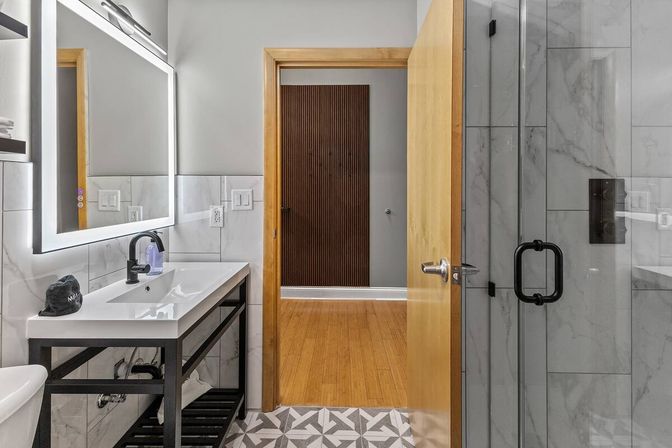 Sleek contemporary bathroom with marble wall tiles, glass shower with black hardware, geometric patterned floor tile, white sink vanity on a black metal frame, illuminated mirror, and an open wooden door leading to a hardwood hallway.
