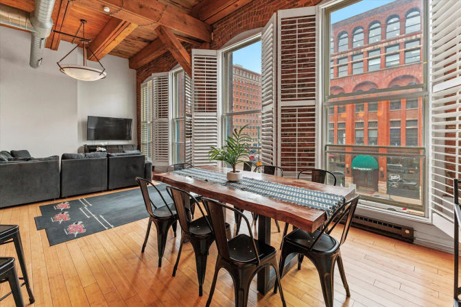 Sunlit industrial downtown loft with exposed wood beams, open living and dining area, live-edge wooden table and black metal chairs, plantation-shutter windows framing red-brick historic buildings outside.