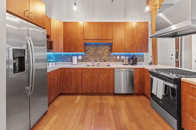 Modern loft-style kitchen with stainless steel appliances, warm wood cabinets, blue LED-lit mosaic backsplash, granite countertops and hardwood floors.