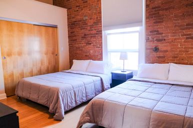 Bright urban loft bedroom with exposed red brick wall, two neatly made beds with neutral bedding, a bedside table and lamp by a sunlit window, hardwood floors and wooden sliding closet doors.
