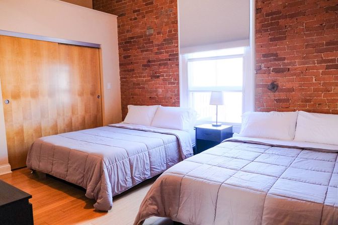 Bright urban loft bedroom with exposed red brick wall, two neatly made beds with neutral bedding, a bedside table and lamp by a sunlit window, hardwood floors and wooden sliding closet doors.