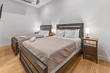 Cozy minimalist guest bedroom with two dark-wood platform beds, beige quilts and white pillows, rolled towels on the beds, wooden nightstands with lamps, and warm hardwood floors.