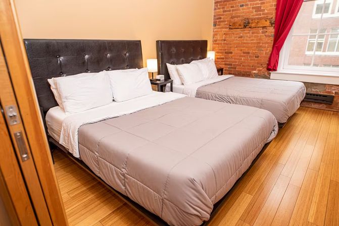 Cozy loft-style guest room with two double beds, tufted dark headboards, exposed brick wall, hardwood floors, bedside lamp and red-curtained window.
