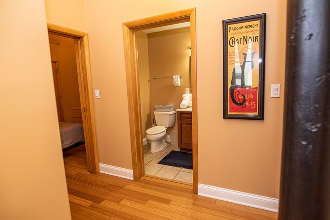 Warm hallway with hardwood floors opening to a small tiled bathroom with a white toilet, wooden vanity, towel and navy bath mat; framed vintage Chat Noir poster on the tan wall and a bedroom doorway to the left.