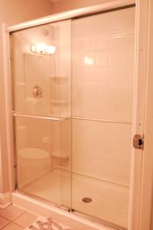 Sliding glass shower doors with chrome towel bar in a white-tiled residential bathroom, showerhead and corner shelf visible