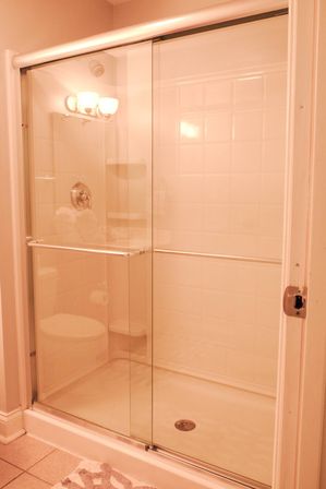 Sliding glass shower doors with chrome towel bar in a white-tiled residential bathroom, showerhead and corner shelf visible