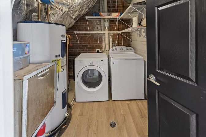Basement laundry room with white front‑load dryer and top‑load washer next to a tall water heater, exposed brick wall, wire shelving, wood‑look vinyl floor and a central floor drain seen through an open black door.