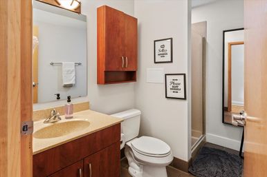 Cozy modern bathroom with wood vanity and wall cabinet, beige countertop, white toilet, glass shower, large mirror, and framed wall signs.
