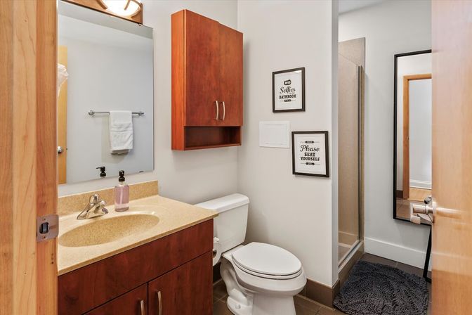 Cozy modern bathroom with wood vanity and wall cabinet, beige countertop, white toilet, glass shower, large mirror, and framed wall signs.