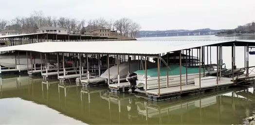 Covered marina with green‑topped pontoon boats docked in slips on a calm lake, shoreline homes and a distant bridge