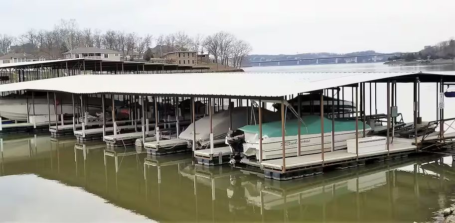 Covered marina with green‑topped pontoon boats docked in slips on a calm lake, shoreline homes and a distant bridge