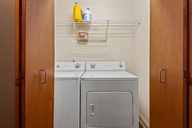 Home laundry closet with side-by-side white washer and dryer, wooden doors, and a wire shelf holding detergent and bleach.