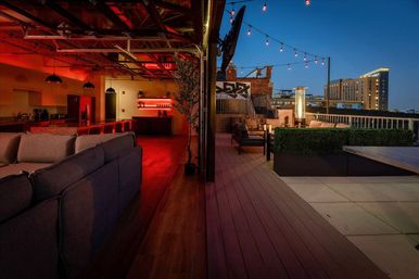 Urban rooftop terrace at dusk — open garage-style lounge with gray sectional, red-lit bar, wood decking, string lights and heated patio seating overlooking a city skyline