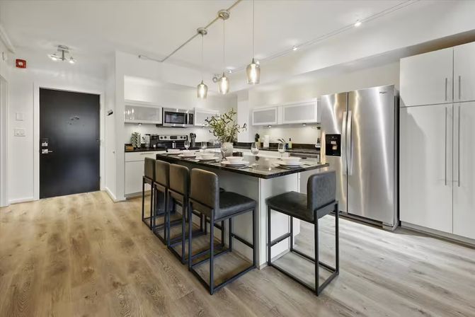 Bright modern open-concept apartment kitchen with black granite island set with five leather bar stools, stainless-steel refrigerator and appliances, white cabinets, pendant lights and light wood flooring.