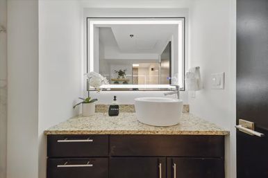 Sleek modern bathroom vanity with granite countertop, round white vessel sink, LED-framed mirror, dark wood cabinets, potted white orchid and soap dispenser.