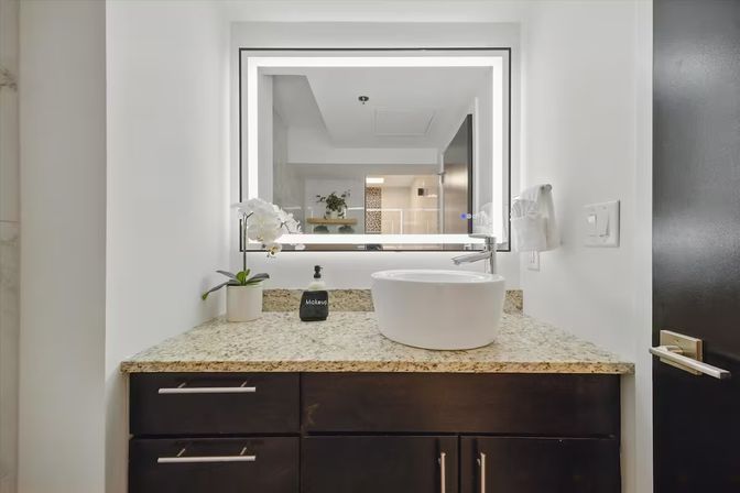 Sleek modern bathroom vanity with granite countertop, round white vessel sink, LED-framed mirror, dark wood cabinets, potted white orchid and soap dispenser.