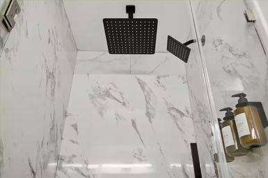 Spa-like marble-tile shower with matte-black square rainfall showerhead and matching handheld, glass door and amber wall-mounted soap dispensers.