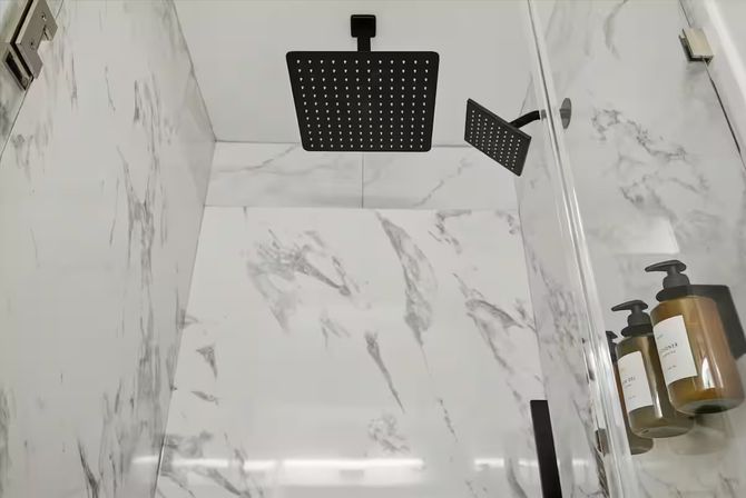 Spa-like marble-tile shower with matte-black square rainfall showerhead and matching handheld, glass door and amber wall-mounted soap dispensers.