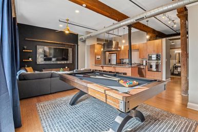 Modern industrial open-concept loft apartment with a wooden pool table, gray sectional sofa, wall-mounted TV, exposed beams and ductwork, hardwood floors, and a stainless-steel kitchen island
